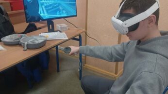 Zdjęcie przedstawia siedzącego chłopca w okularach VR, który trzyma w dłoniach kontrolery. Przed nim na biurku stoi monitor wyświetlający widok z gry lub aplikacji osadzonej w środowisku miejskim. W tle widoczne są tablice edukacyjne zawieszone na ścianie sali lekcyjnej.