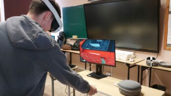 Zdjęcie przedstawia młodego mężczyznę w szarej bluzie z kapturem, który korzysta z gogli VR i kontrolera w sali lekcyjnej. Stoi on tyłem do obiektywu, patrząc w stronę monitora, na którym wyświetlany jest widok z symulacji — czerwone drzwi samochodu. W tle widoczna jest zielona tablica z napisem oraz duży ekran interaktywny. Na biurku obok monitora leży etui na sprzęt oraz inne akcesoria elektroniczne.