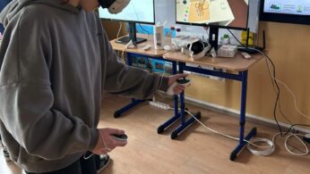 Dziewczyna w okularach VR i z kontrolerami w dłoniach uczestniczy w symulacji zawodowej w klasie. Na monitorze przed nią widać podgląd aplikacji, która przedstawia instruktaż medyczny lub laboratoryjny. W tle znajduje się biała tablica oraz drugi monitor. Obok stoi inna osoba, również korzystająca z zestawu do rzeczywistości wirtualnej.