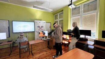 Zdjęcie przedstawia nowoczesną salę lekcyjną z jasnozielonymi ścianami, w której dwoje uczniów korzysta z gogli VR. Chłopak stoi na środku pokoju i używa kontrolerów, podczas gdy dziewczyna siedzi tyłem przy biurku przed dużym monitorem. Obok nich stoi kobieta w czarnej kurtce, która obserwuje ich działania. W tle widać baner z napisem „Nauka przez całe życie” oraz okno z zaciągniętymi roletami.