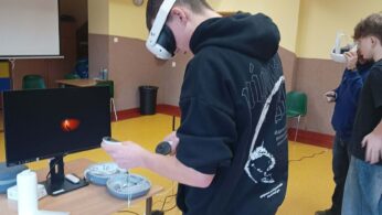 Młody mężczyzna w czarnej bluzie z kapturem stoi w sali lekcyjnej, korzystając z gogli VR i kontrolerów. Przed nim na biurku znajduje się monitor wyświetlający ciemny obraz z jasnym punktem. W tle widoczne są inne osoby oraz wyposażenie sali, w tym projektor pod sufitem.