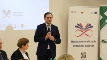 Zbliżenie na mężczyznę w okularach i granatowym garniturze, który przemawia do mikrofonu podczas konferencji. Obok niego przy stole siedzi kobieta w czarnym żakiecie i białej koszuli. W tle widoczny jest ekran z napisem dotyczącym debaty o edukacji oraz roll-up z logo projektu.
