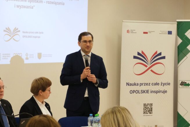Zbliżenie na mężczyznę w okularach i granatowym garniturze, który przemawia do mikrofonu podczas konferencji. Obok niego przy stole siedzi kobieta w czarnym żakiecie i białej koszuli. W tle widoczny jest ekran z napisem dotyczącym debaty o edukacji oraz roll-up z logo projektu.