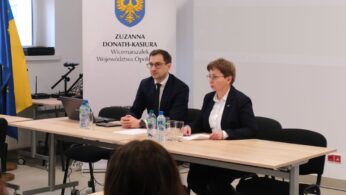Mężczyzna i kobieta w okularach siedzą przy jasnym stole konferencyjnym na tle białej ścianki z herbem i napisem 
