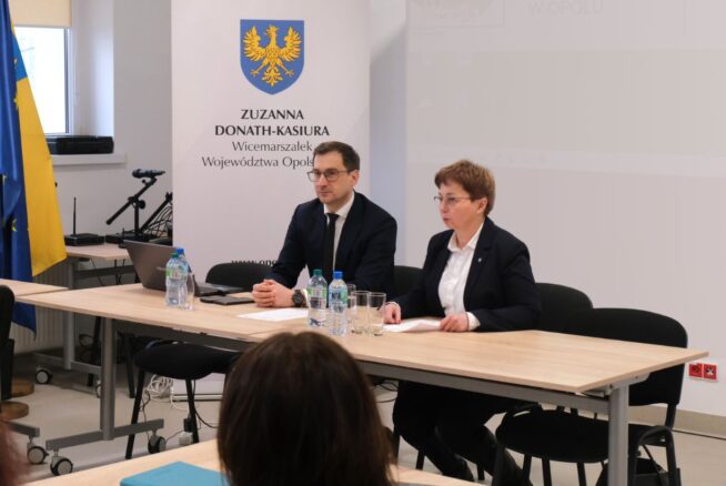 Mężczyzna i kobieta w okularach siedzą przy jasnym stole konferencyjnym na tle białej ścianki z herbem i napisem "Zuzanna Donath-Kasiura". Na stole znajdują się butelki wody, szklanki oraz laptop. W tle widać fragmenty flagi Polski i regionu.