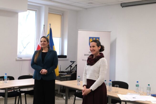 Dwie uśmiechnięte kobiety stoją w sali konferencyjnej obok flag i roll-upu z herbem województwa opolskiego. Jedna z nich ma na sobie zielony żakiet, a druga biały sweter i bordową spódnicę. W tle widać stoły konferencyjne z butelkami wody i dokumentami. Pomieszczenie jest jasne, oświetlone światłem dziennym wpadającym przez okno.
