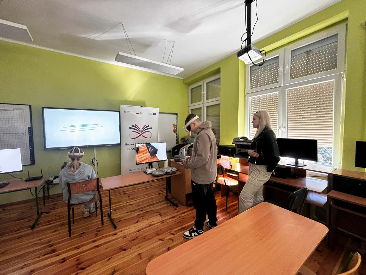Zdjęcie przedstawia nowoczesną salę lekcyjną z jasnozielonymi ścianami, w której dwoje uczniów korzysta z gogli VR. Chłopak stoi na środku pokoju i używa kontrolerów, podczas gdy dziewczyna siedzi tyłem przy biurku przed dużym monitorem. Obok nich stoi kobieta w czarnej kurtce, która obserwuje ich działania. W tle widać baner z napisem „Nauka przez całe życie” oraz okno z zaciągniętymi roletami.