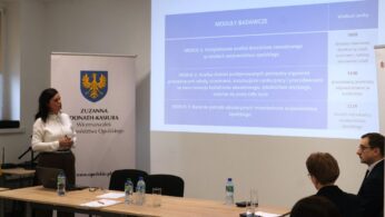 Kobieta stoi przy ekranie projekcyjnym, na którym wyświetlany jest slajd z napisem „ZAKRES BADAŃ” i tabelą dotyczącą modułów badawczych. Po prawej stronie, tyłem do obiektywu, siedzą uczestnicy spotkania wsłuchani w prezentację. Obok ekranu stoi pionowy baner z herbem województwa opolskiego.
