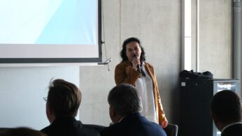 Kobieta w brązowej marynarce i białej koszuli przemawia do mikrofonu, patrząc w stronę publiczności. Stoi obok ekranu projekcyjnego w minimalistycznym, betonowym wnętrzu. Przed nią widoczne są tyły głów uczestników konferencji słuchających wypowiedzi.