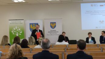Szerokie ujęcie sali konferencyjnej, w której trwa debata na temat wdrażania projektów kształcenia zawodowego. Mężczyzna w garniturze stoi przy mównicy z czerwonymi aktami i przemawia do mikrofonu. W tle widoczne są banery instytucji regionalnych oraz ekran z prezentacją. Na pierwszym planie widać tyły głów uczestników siedzących w ławkach.