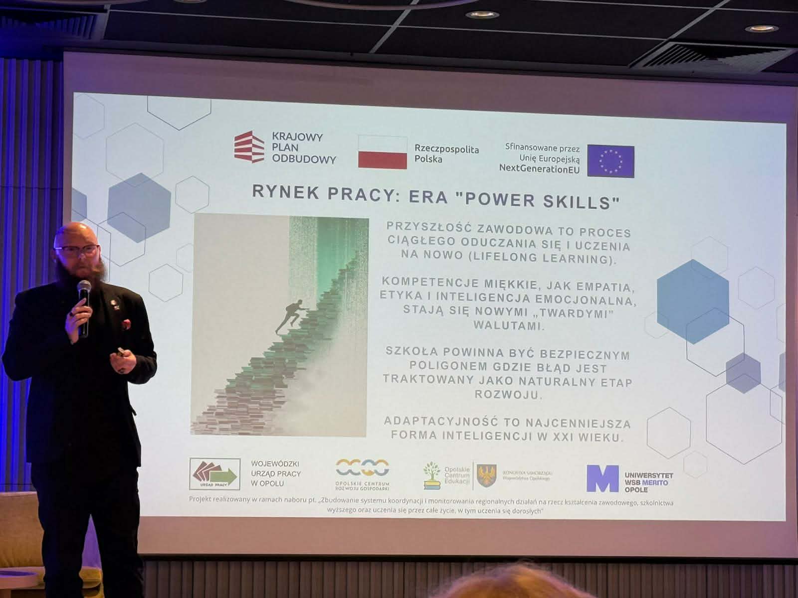 Prelegent z brodą i w okularach stoi przy dużym ekranie, na którym widnieje napis „RYNEK PRACY: ERA 'POWER SKILLS'”. Slajd zawiera tekst dotyczący kompetencji miękkich i uczenia się przez całe życie (lifelong learning). Na dole ekranu widoczne są logotypy instytucji unijnych i regionalnych.