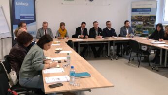 Szerokie ujęcie sali konferencyjnej, w której uczestnicy siedzą przy stołach ustawionych w kształt litery U. Na pierwszym planie widać kobietę robiącą notatki, a w tle ściany z logotypami urzędowymi oraz baner „Fundusze Europejskie dla opolskiego”. Atmosfera wskazuje na oficjalne spotkanie robocze lub szkolenie.