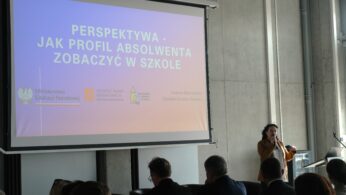 Prelegentka przemawia do publiczności zgromadzonej w nowoczesnej sali konferencyjnej. W tle widoczny jest duży ekran z prezentacją zatytułowaną „Perspektywa – jak profil absolwenta zobaczyć w szkole”. Na dole slajdu widnieją logotypy Ministerstwa Edukacji Narodowej oraz innych instytucji edukacyjnych.