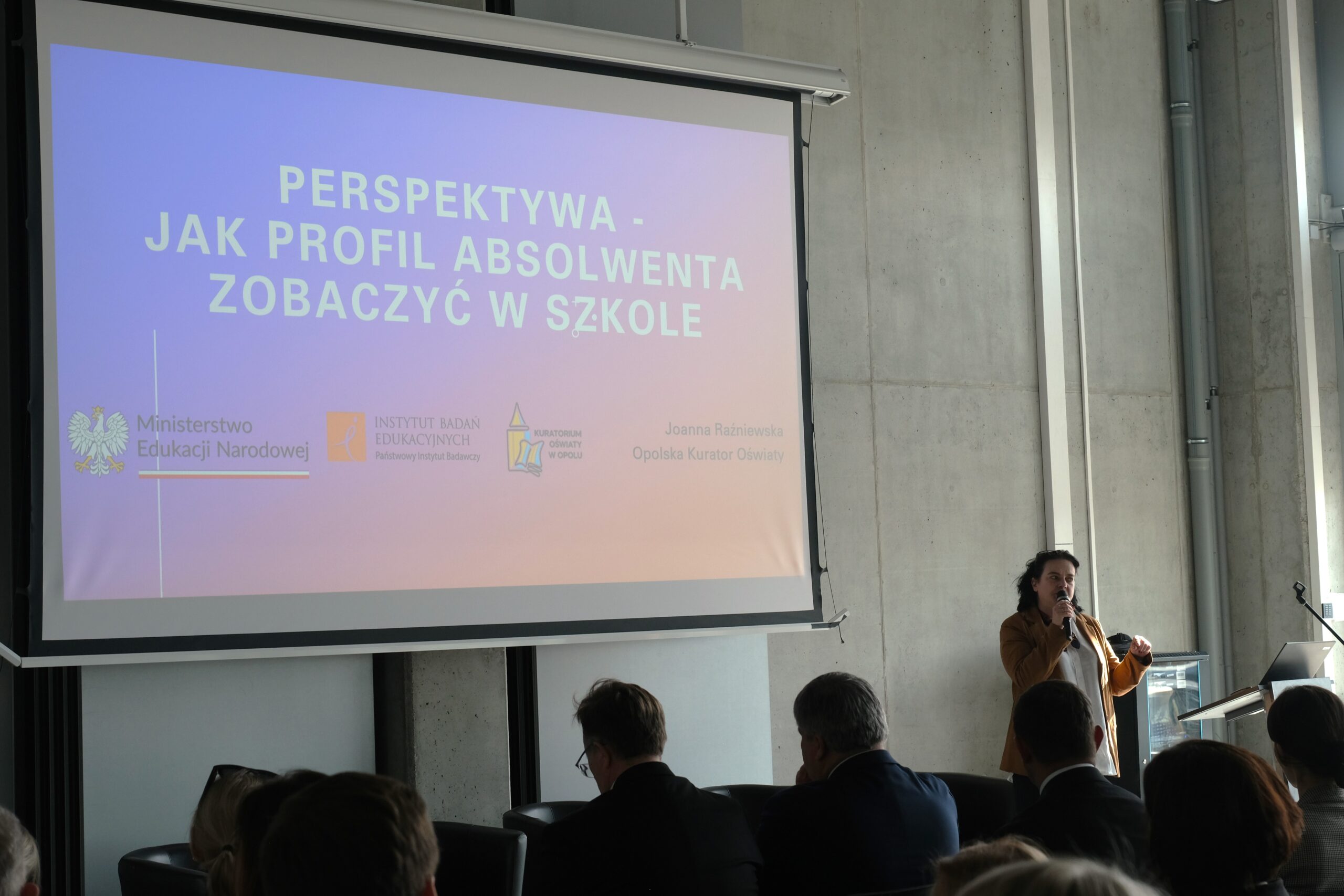 Prelegentka przemawia do publiczności zgromadzonej w nowoczesnej sali konferencyjnej. W tle widoczny jest duży ekran z prezentacją zatytułowaną „Perspektywa – jak profil absolwenta zobaczyć w szkole”. Na dole slajdu widnieją logotypy Ministerstwa Edukacji Narodowej oraz innych instytucji edukacyjnych.