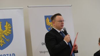 Bliskie ujęcie mężczyzny w okularach i ciemnym garniturze, który przemawia do mikrofonu podczas oficjalnego spotkania. W dłoni trzyma czerwoną teczkę z dokumentami. W tle widoczny jest fragment białego baneru z herbem województwa opolskiego (żółty orzeł na błękitnym polu). Postać jest zwrócona lekko w prawą stronę.