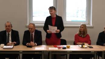 Kobieta w czerwonej sukience i czarnym żakiecie stoi przy stole konferencyjnym, trzymając w dłoniach dokumenty i przemawiając do zebranych. Po jej obu stronach siedzą mężczyźni i kobieta w strojach biznesowych, słuchając wystąpienia. W tle widoczne są duże okna, przez które wpada dzienne światło.