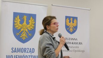 Kobieta w okularach o różowych oprawkach i szarym swetrze przemawia do mikrofonu, stojąc przed banerami informacyjnymi. Ma spięte włosy i gestykuluje lewą ręką, w której trzyma pilot do prezentacji. W tle widnieje herb regionu oraz napis „Zuzanna Donath-Kasiura – Wicemarszałek Województwa Opolskiego”. Zdjęcie przedstawia profil prelegentki.