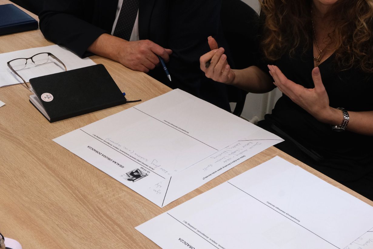 Zdjęcie przedstawia dłonie i sylwetki osób uczestniczących w warsztatach, siedzących przy stole z arkuszami papieru. Jedna z osób gestykuluje dłońmi, co sugeruje prowadzenie ożywionej rozmowy lub wyjaśnianie koncepcji. Na pierwszym planie leży wypełniony arkusz „IDEALNA USŁUGA DORADCZA” z naniesionymi odręcznymi zapiskami. Obok widoczny jest czarny notatnik oraz okulary korekcyjne.