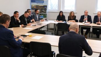 Grupa osób siedząca przy długim stole konferencyjnym, widoczna z perspektywy jednego z uczestników (widoczne plecy osoby na pierwszym planie). Uczestnicy spotkania mają przed sobą filiżanki z kawą, szklanki z sokiem oraz materiały szkoleniowe. Scena przedstawia moment uważnego słuchania podczas narady.
