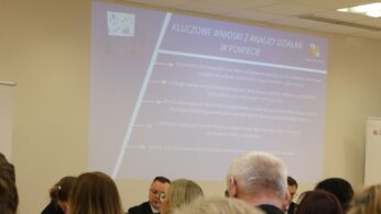 Zbliżenie na slajd prezentacji multimedialnej wyświetlanej na białej ścianie podczas debaty. Tekst na slajdzie dotyczy wniosków z analizy działań wspierających kształcenie zawodowe w powiecie prudnickim. W dolnej części kadru widać rozmazane sylwetki osób siedzących w sali konferencyjnej. Na slajdzie widoczny jest również herb powiatu.