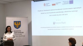 Zuzanna Donath-Kasiura, Wicemarszałek Województwa Opolskiego, stoi obok ekranu projekcyjnego i przemawia do publiczności. Na ekranie wyświetlany jest tytuł projektu dotyczącego koordynacji działań na rzecz kształcenia zawodowego i uczenia się przez całe życie. Obok niej znajduje się roll-up z herbem województwa opolskiego.