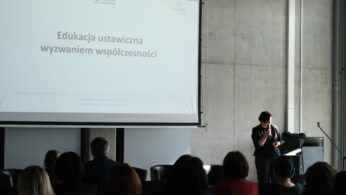 Prelegentka stoi obok ekranu, na którym wyświetlany jest slajd o treści „Edukacja ustawiczna wyzwaniem współczesności”. W górnym rogu ekranu widoczne jest logo Politechniki Opolskiej. Sala jest wypełniona słuchaczami, co podkreśla edukacyjny charakter spotkania.