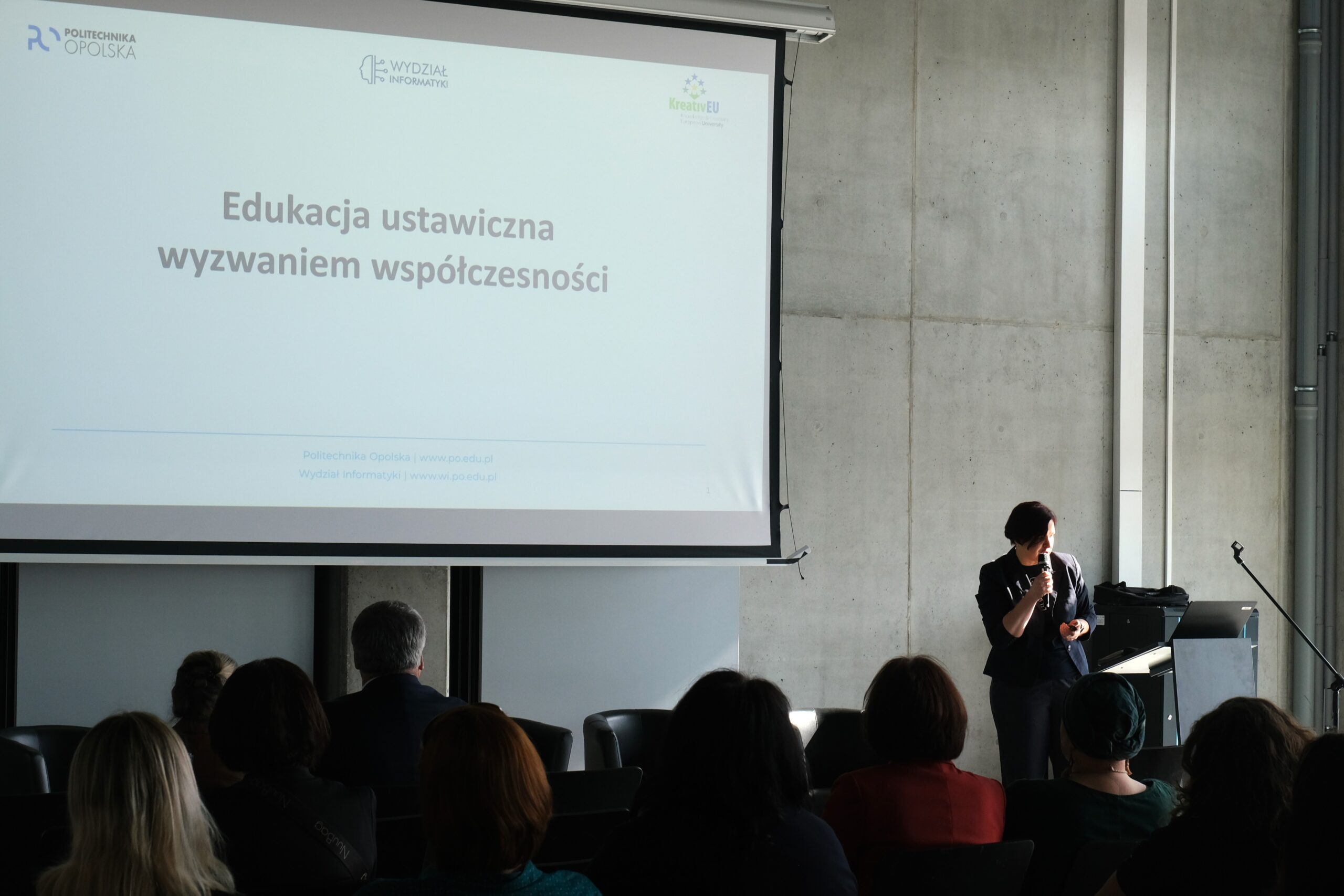 Prelegentka stoi obok ekranu, na którym wyświetlany jest slajd o treści „Edukacja ustawiczna wyzwaniem współczesności”. W górnym rogu ekranu widoczne jest logo Politechniki Opolskiej. Sala jest wypełniona słuchaczami, co podkreśla edukacyjny charakter spotkania.