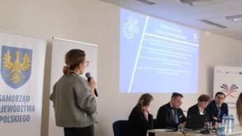 Kobieta prowadząca prezentację stoi bokiem do obiektywu, trzymając mikrofon i patrząc w stronę ekranu. Na ścianie wyświetlany jest niebieski slajd z nagłówkiem „Wnioski dotyczące kształcenia zawodowego” i ikoną klucza oraz zębatki. Przy stole obok siedzą trzej eksperci śledzący przebieg wystąpienia. Po lewej stronie kadru widoczny jest duży baner Samorządu Województwa Opolskiego.