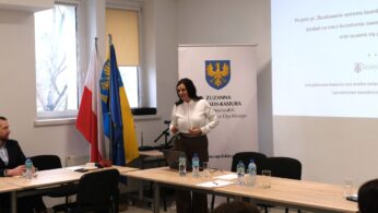 Wicemarszałek Zuzanna Donath-Kasiura stoi przy ekranie prezentacyjnym w jasnej sali, przygotowując się do dalszej części wystąpienia. Obok niej widoczne są flagi Polski oraz województwa opolskiego ustawione na stojakach. Na ekranie wciąż widoczny jest slajd tytułowy dotyczący badań i analiz rynku pracy.