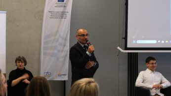 Mężczyzna w okularach i garniturze przemawia do mikrofonu, stojąc obok białego baneru z flagą Unii Europejskiej. Po lewej stronie widoczna jest tłumaczka języka migowego, która przekłada wystąpienie. W tle siedzi młody mężczyzna w białej koszuli, przysłuchując się debacie.