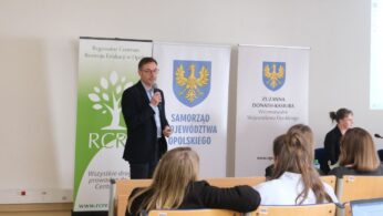 Mężczyzna w okularach i ciemnej marynarce stoi na środku, przemawiając do mikrofonu podczas spotkania edukacyjnego. Po jego prawej stronie znajdują się trzy pionowe banery reklamowe z logo RCRE oraz herbem województwa. W tle po prawej stronie siedzą dwie kobiety przy stole z laptopem. Widok z perspektywy uczestnika siedzącego w ostatnich rzędach sali.