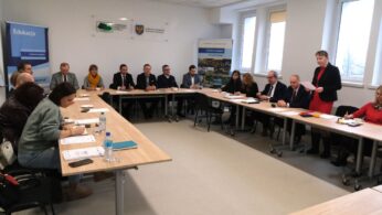 Szeroki kadr na całą salę konferencyjną, w której kobieta w czerwonym stroju stoi i odczytuje treść z kartki. Pozostali uczestnicy, siedzący wzdłuż stołów, obserwują mówczynię lub sporządzają notatki w swoich materiałach. Pomieszczenie jest jasne, o biurowym wystroju, z widocznymi elementami identyfikacji wizualnej funduszy unijnych.