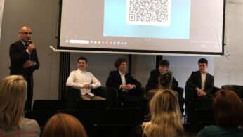 Szerokie ujęcie sceny, na której czterech młodych mężczyzn w eleganckich strojach siedzi w fotelach podczas panelu dyskusyjnego. Nad nimi znajduje się ekran wyświetlający duży kod QR. Po lewej stronie stoi moderator z mikrofonem, a na pierwszym planie widać publiczność.