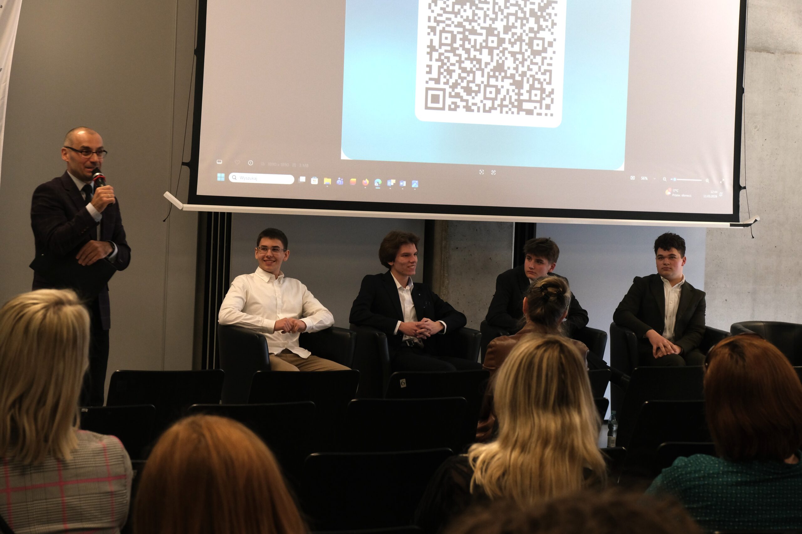 Szerokie ujęcie sceny, na której czterech młodych mężczyzn w eleganckich strojach siedzi w fotelach podczas panelu dyskusyjnego. Nad nimi znajduje się ekran wyświetlający duży kod QR. Po lewej stronie stoi moderator z mikrofonem, a na pierwszym planie widać publiczność.