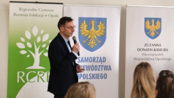 Portretowe ujęcie mężczyzny w średnim wieku, ubranego w ciemną marynarkę i niebieską koszulę, który wygłasza przemówienie. W jednej ręce trzyma mikrofon, a w drugiej pilot do przełączania slajdów. Stoi na tle białych ścian i banerów z oficjalnymi oznaczeniami województwa opolskiego. Postać jest skupiona i patrzy przed siebie na słuchaczy.
