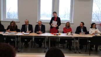 Zbliżenie na centralną część stołu konferencyjnego, przy którym stoi kobieta prowadząca prezentację. Pozostali uczestnicy siedzą rzędem, patrząc w stronę obiektywu lub na mówczynię, a przed nimi leżą dokumenty i drobne poczęstunki. Zdjęcie dokumentuje przebieg oficjalnego panelu dyskusyjnego.