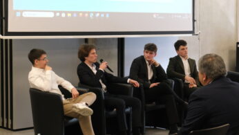 Uczestnicy panelu dyskusyjnego siedzą w czarnych skórzanych fotelach pod ekranem projekcyjnym. Jeden z młodych mężczyzn trzyma mikrofon i zabiera głos, podczas gdy pozostali słuchają. Atmosfera spotkania wydaje się profesjonalna i sprzyjająca wymianie opinii.