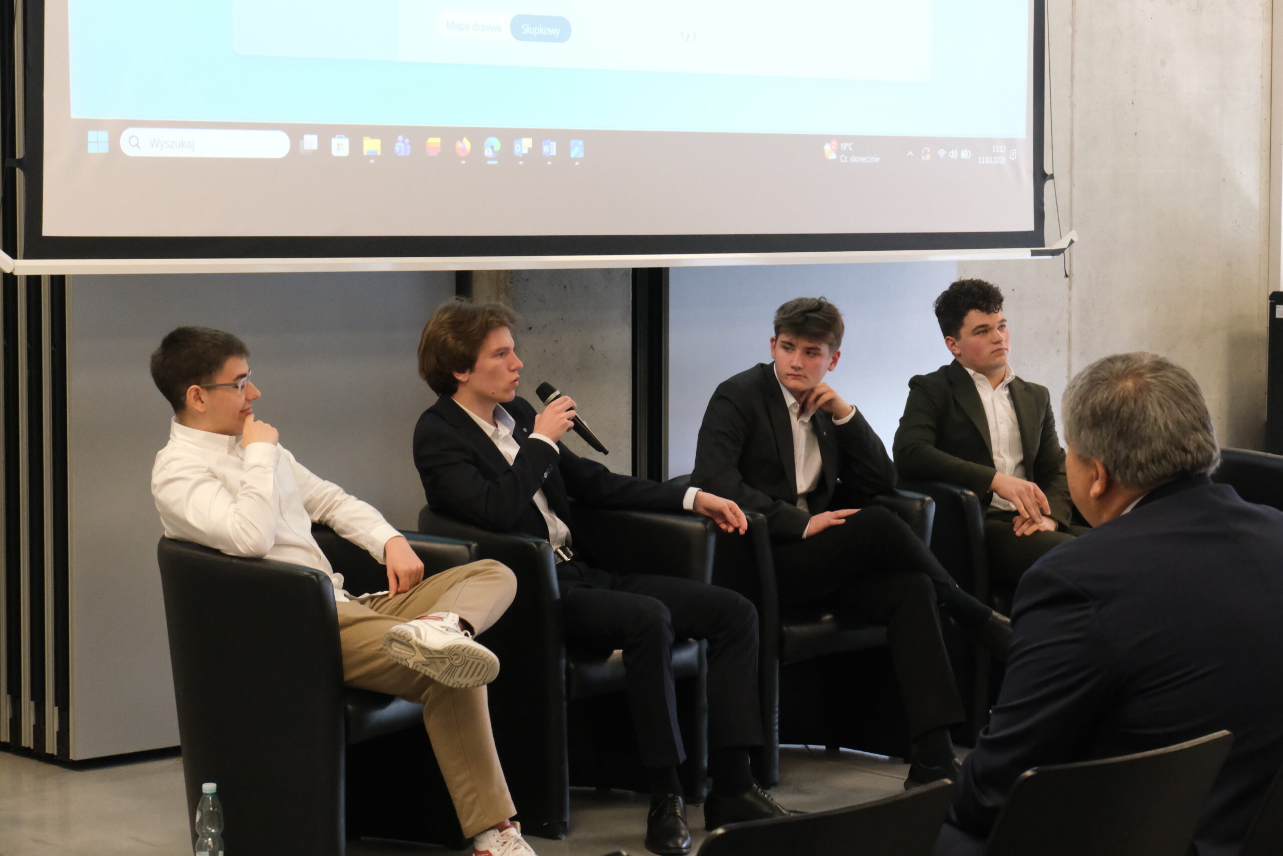 Uczestnicy panelu dyskusyjnego siedzą w czarnych skórzanych fotelach pod ekranem projekcyjnym. Jeden z młodych mężczyzn trzyma mikrofon i zabiera głos, podczas gdy pozostali słuchają. Atmosfera spotkania wydaje się profesjonalna i sprzyjająca wymianie opinii.