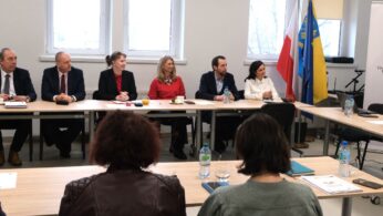 Grupa sześciu osób siedzi przy głównym stole konferencyjnym, uśmiechając się lub patrząc w stronę prezentera poza kadrem. Na stole widać szklankę soku, filiżanki i dokumenty, a w tle znajdują się flagi oraz okna. Zdjęcie oddaje profesjonalną, a zarazem przyjazną atmosferę spotkania.