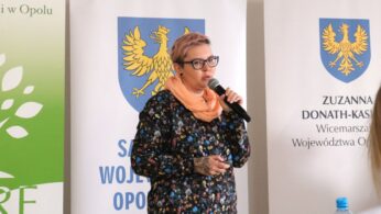 Kobieta z krótkimi, różowymi włosami i w okularach mówi do mikrofonu, trzymając w drugiej ręce pilot do slajdów. Jest ubrana w ciemną bluzkę w kolorowe kwiaty i jasny, brzoskwiniowy szal. Na dłoni prelegentki widoczny jest tatuaż. W tle znajdują się banery z logo Regionalnego Centrum Rozwoju Edukacji oraz herbem regionu.