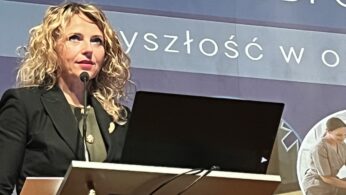 Zbliżenie na kobietę o blond włosach przemawiającą do mikrofonu przy mównicy z laptopem. Nad nią widnieje fragment baneru z napisem „ZAWODY ODPO...”. W dolnym rogu widoczny jest logotyp Krajowego Planu Odbudowy.