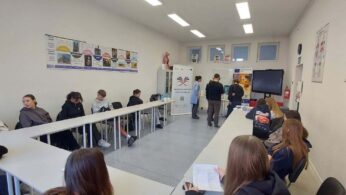 Szerokie ujęcie sali wykładowej wypełnionej młodzieżą. Uczniowie siedzą przy białych stołach ustawionych w kształcie litery U. Na ścianach wiszą plakaty edukacyjne dotyczące epok historycznych oraz anatomii człowieka. W centralnej części sali dwie osoby dorosłe (kobieta w niebieskim fartuchu i mężczyzna w czarnej koszulce) przygotowują stanowisko z technologią VR. W tle widać banery informacyjne oraz duży ekran interaktywny.