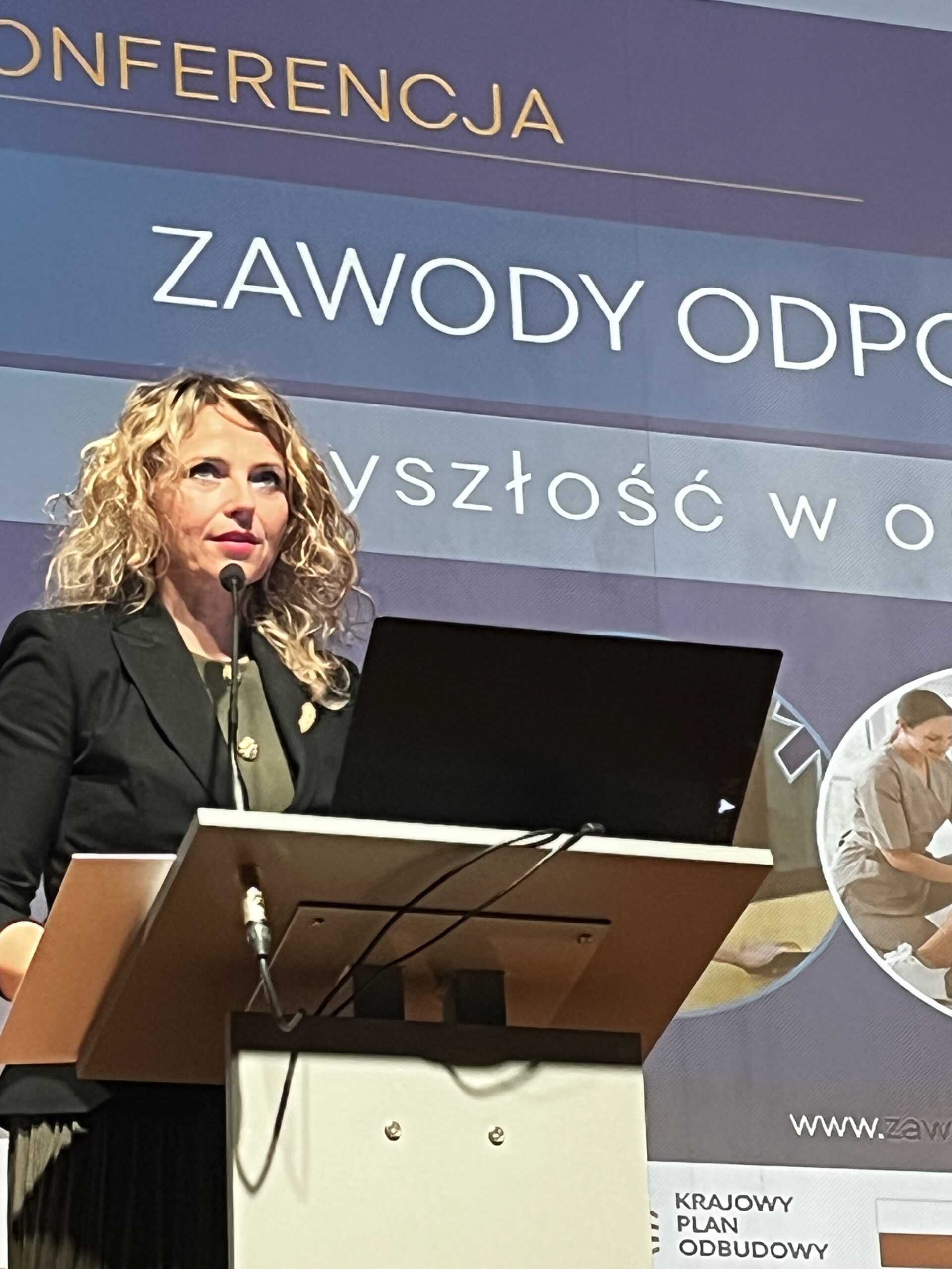 Zbliżenie na kobietę o blond włosach przemawiającą do mikrofonu przy mównicy z laptopem. Nad nią widnieje fragment baneru z napisem „ZAWODY ODPO...”. W dolnym rogu widoczny jest logotyp Krajowego Planu Odbudowy.