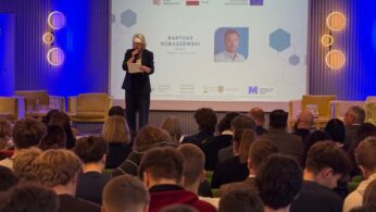 Prelegentka w okularach stoi na scenie i czyta z kartki, a na ekranie za nią wyświetla się slajd z nazwiskiem „BARTOSZ ROBASZEWSKI” i jego zdjęciem. Sala jest wypełniona słuchaczami, których widać od tyłu. Wnętrze jest oświetlone ciepłym światłem z ozdobnych lamp ściennych.