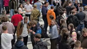 Wysokie, nowoczesne wnętrze hali wypełnione tłumem ludzi uczestniczących w targach pracy lub wydarzeniu edukacyjnym. Wzdłuż przeszklonej ściany ustawione są stoiska wystawiennicze z banerami, między innymi firmy GruppoRE. Uczestnicy spacerują między stanowiskami po charakterystycznej podłodze w biało-czarną szachownicę.