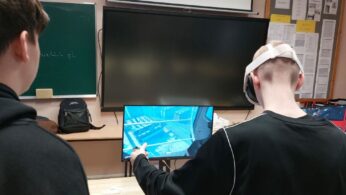 Fotografia ukazuje dwóch młodych mężczyzn od tyłu, obserwujących symulację na monitorze komputera w klasie. Jeden z nich ma na głowie biały zestaw VR i trzyma w dłoni kontroler, kierując go w stronę ekranu. Na monitorze widoczny jest widok z perspektywy pierwszej osoby, przedstawiający miejską infrastrukturę lub plac budowy w niebieskawej kolorystyce. W tle widać tablicę szkolną, wyłączony monitor ścienny oraz gazetkę ścienną z ogłoszeniami.