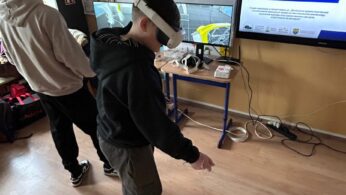 Dwóch chłopców korzysta z gogli VR w sali lekcyjnej, trzymając w dłoniach bezprzewodowe kontrolery. Na ekranach monitorów wyświetlany jest podgląd środowiska 3D, przedstawiający halę warsztatową lub magazynową. Na ścianie wisi duży ekran z prezentacją o wsparciu nowoczesnego kształcenia zawodowego. Nad tablicą widoczny jest polski godło oraz krzyż.