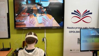 Zbliżenie na ucznia siedzącego tyłem do aparatu w białych goglach VR, który patrzy na duży interaktywny ekran. Na wyświetlaczu widać widok z perspektywy pierwszej osoby, przedstawiający ręce w wirtualnej rzeczywistości operujące nad biurkiem z laptopem i notatkami. Po prawej stronie stoi biały baner informacyjny z logotypami Unii Europejskiej i programów regionalnych. Na stole obok leży dodatkowy zestaw gogli i akcesoria.