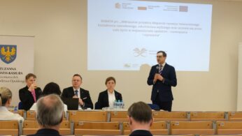 Szerokie ujęcie sali wykładowej, w której trwa debata. Na podium przy stole siedzą trzy osoby, a czwarta — mężczyzna w garniturze — stoi i przemawia do mikrofonu. W tle znajduje się duży ekran z tytułem wydarzenia, a na pierwszym planie widoczne są tyły głów uczestników siedzących w ławkach.