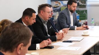 Grupa mężczyzn w garniturach siedzi przy długim stole, uczestnicząc w dyskusji lub konferencji. Na pierwszym planie widać dokumenty, telefony komórkowe oraz filiżankę kawy. W tle znajduje się kolorowy baner z grafikami przedstawiającymi infrastrukturę i obiekty użyteczności publicznej.